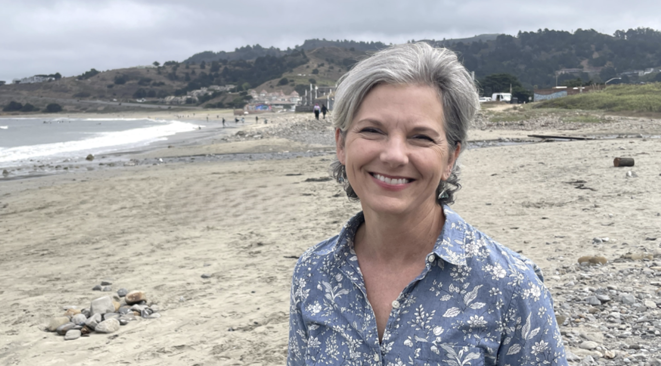 Margo Meiman smiling on a Pacifica beach with the shoreline and coastal hills behind her