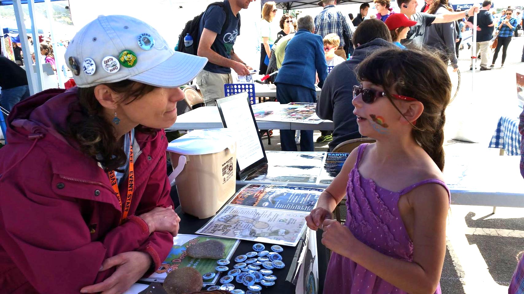 Margo, wearing an Earth Hero cap and event staff lanyard, talking with a young girl at an outdoor Earth Day festival booth in Pacifica