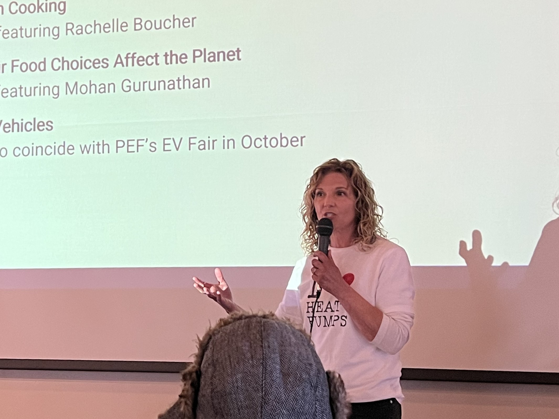 Margo speaking into a microphone at a climate event, wearing an I heart heat pumps shirt in front of a presentation slide